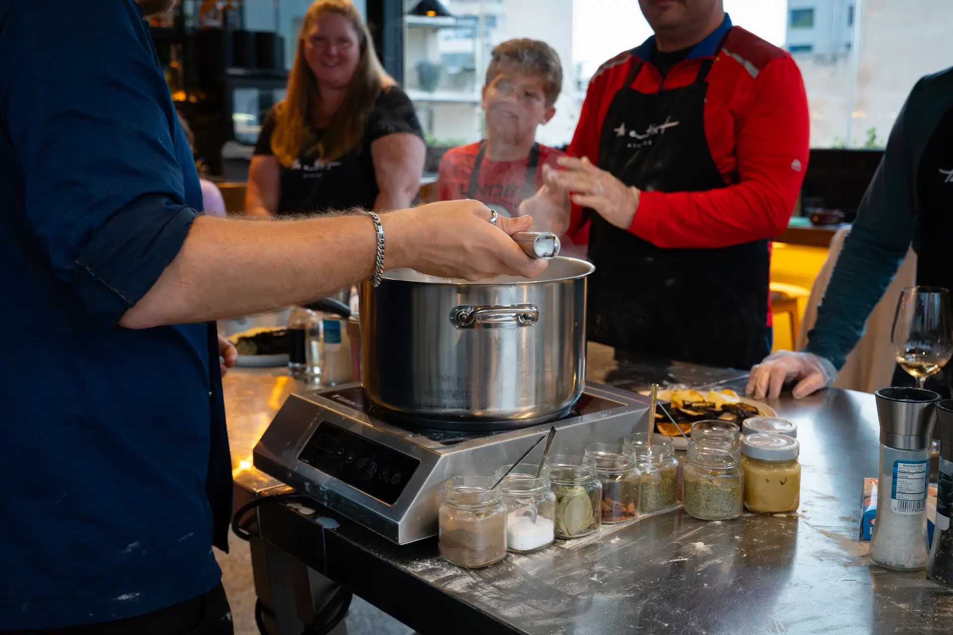 Chef engaging the group around a steaming pot — spices lined up, full attention